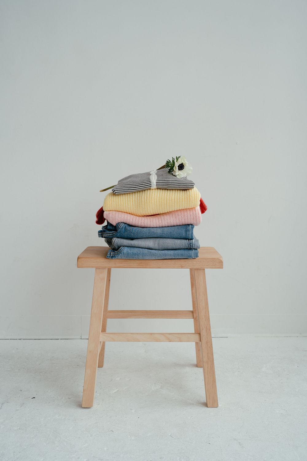 A neatly folded stack of colorful clothes in pink, yellow, gray, and denim sits on a wooden stool with a flower on top.