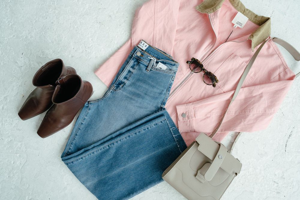A flat lay of a pink shirt, blue jeans, brown ankle boots, sunglasses, and a gray crossbody bag on a light surface.