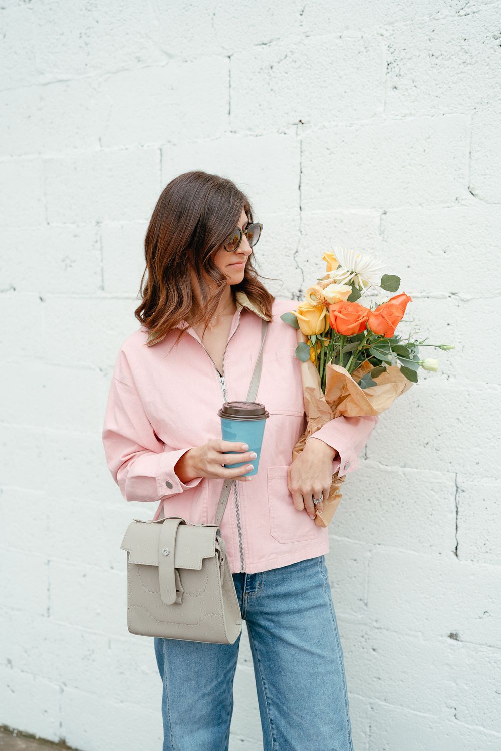 A stylish woman in a pink jacket and jeans holds a bouquet of orange roses and a coffee cup against a white brick wall.