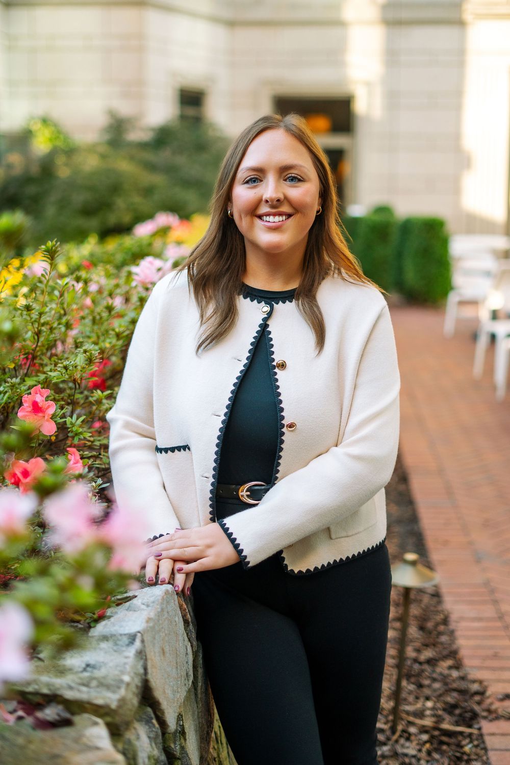 A smiling young woman in a white jacket poses beside colorful flowers in a garden.
