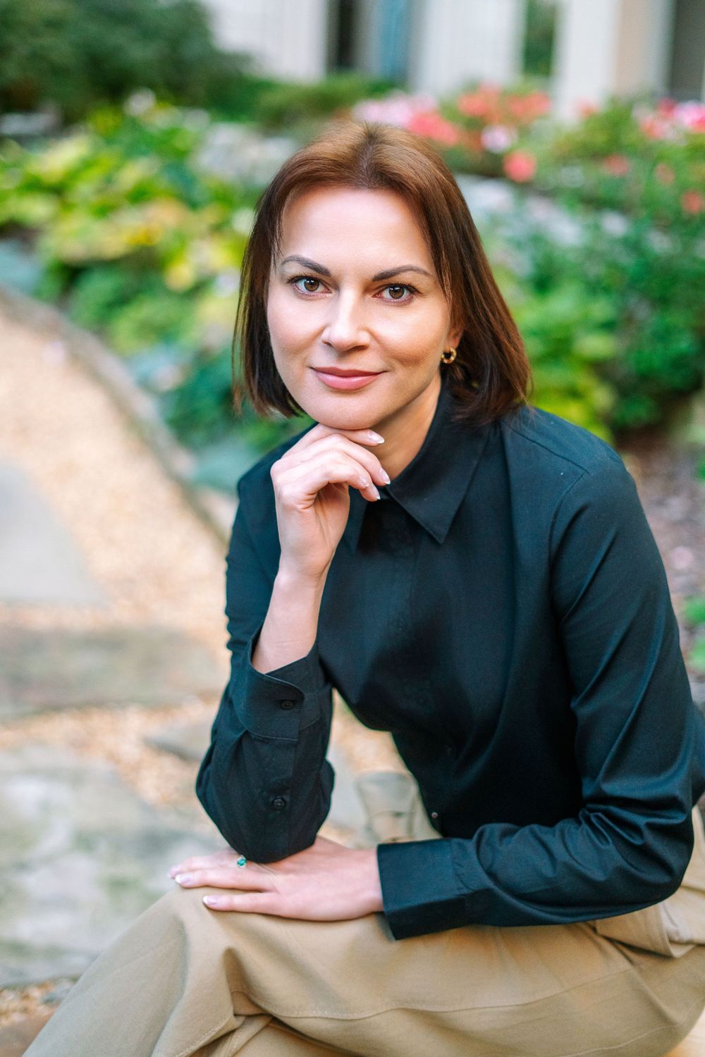 Professional woman in black blouse posing thoughtfully in a green garden courtyard.