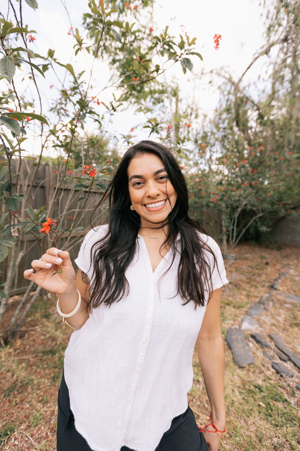 A smiling woman with long dark hair in a white linen shirt stands in a garden holding a small orange flower.