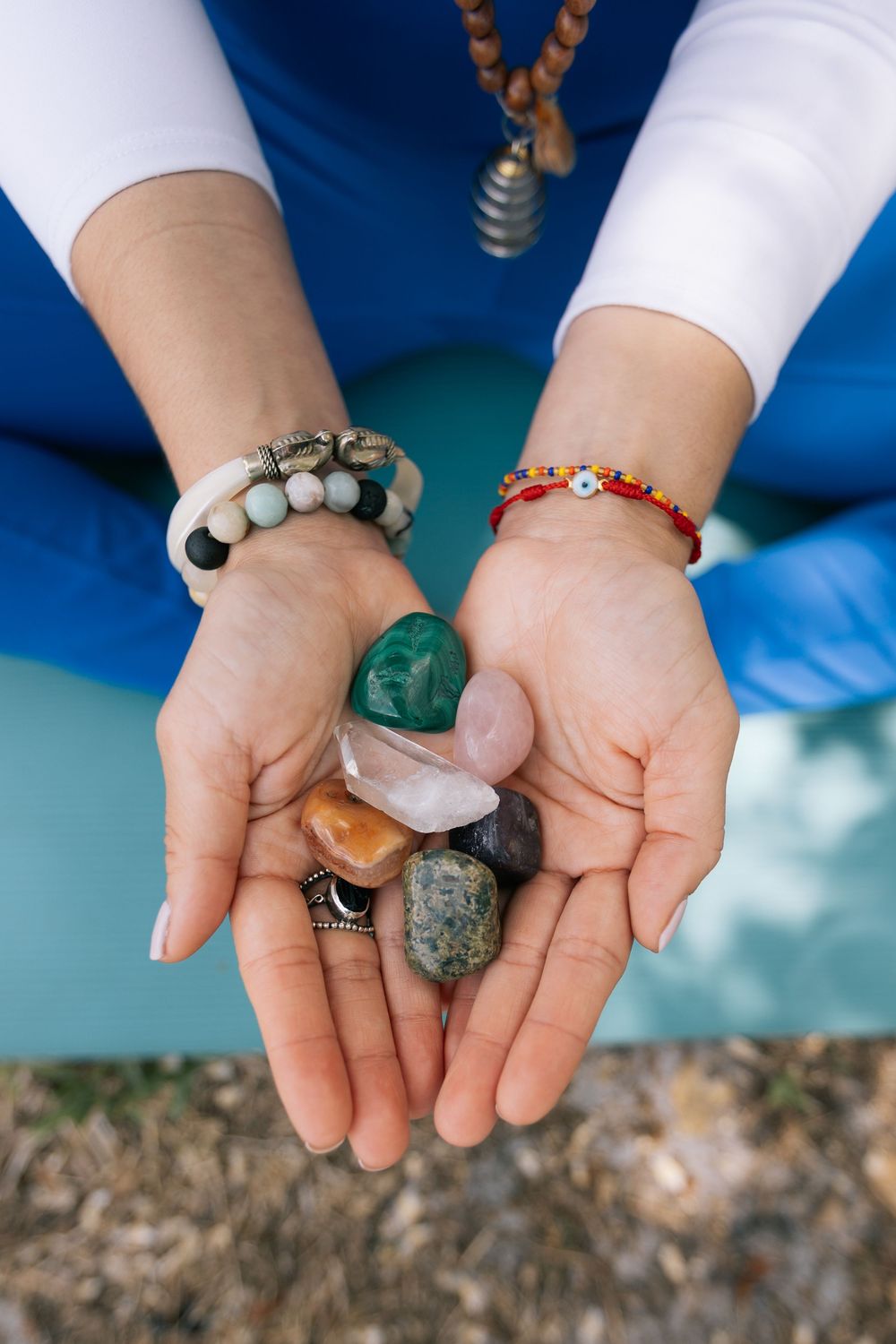 Open hands hold a collection of colorful healing crystals and gemstones, with beaded bracelets and a mala necklace visible.