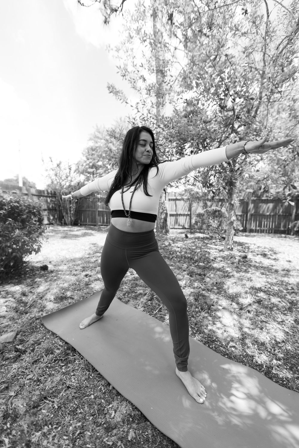 A black and white photo of a woman in activewear practicing a warrior yoga pose on a mat in a backyard.