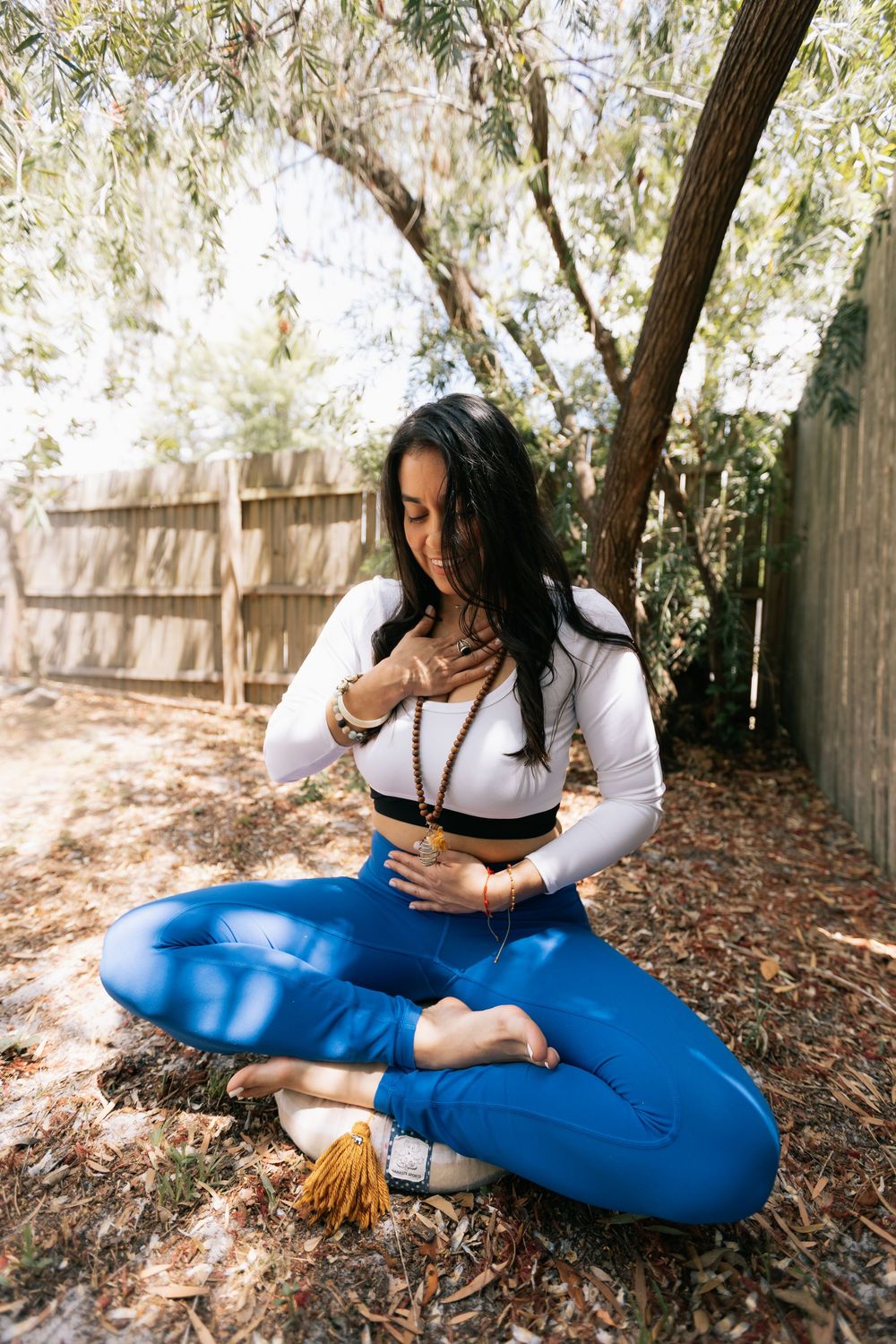 Woman in blue leggings sits cross-legged under a tree, hand on her heart in a calm outdoor meditation pose.