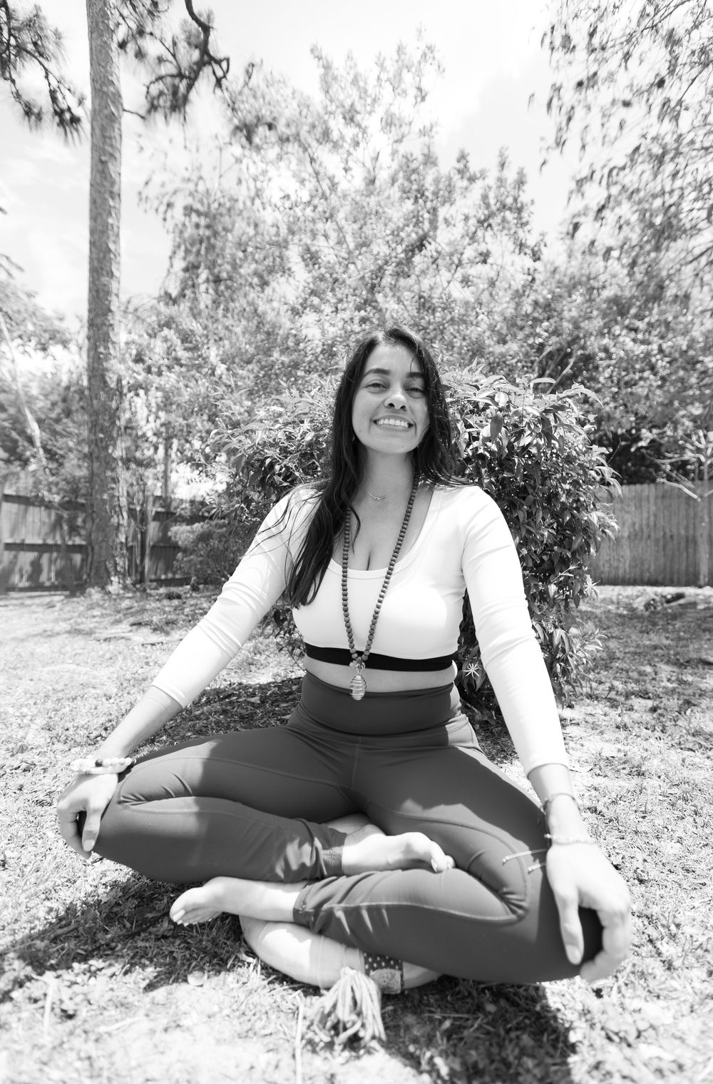 A black and white photo of a smiling woman in a long beaded necklace meditating cross-legged on grass outdoors.
