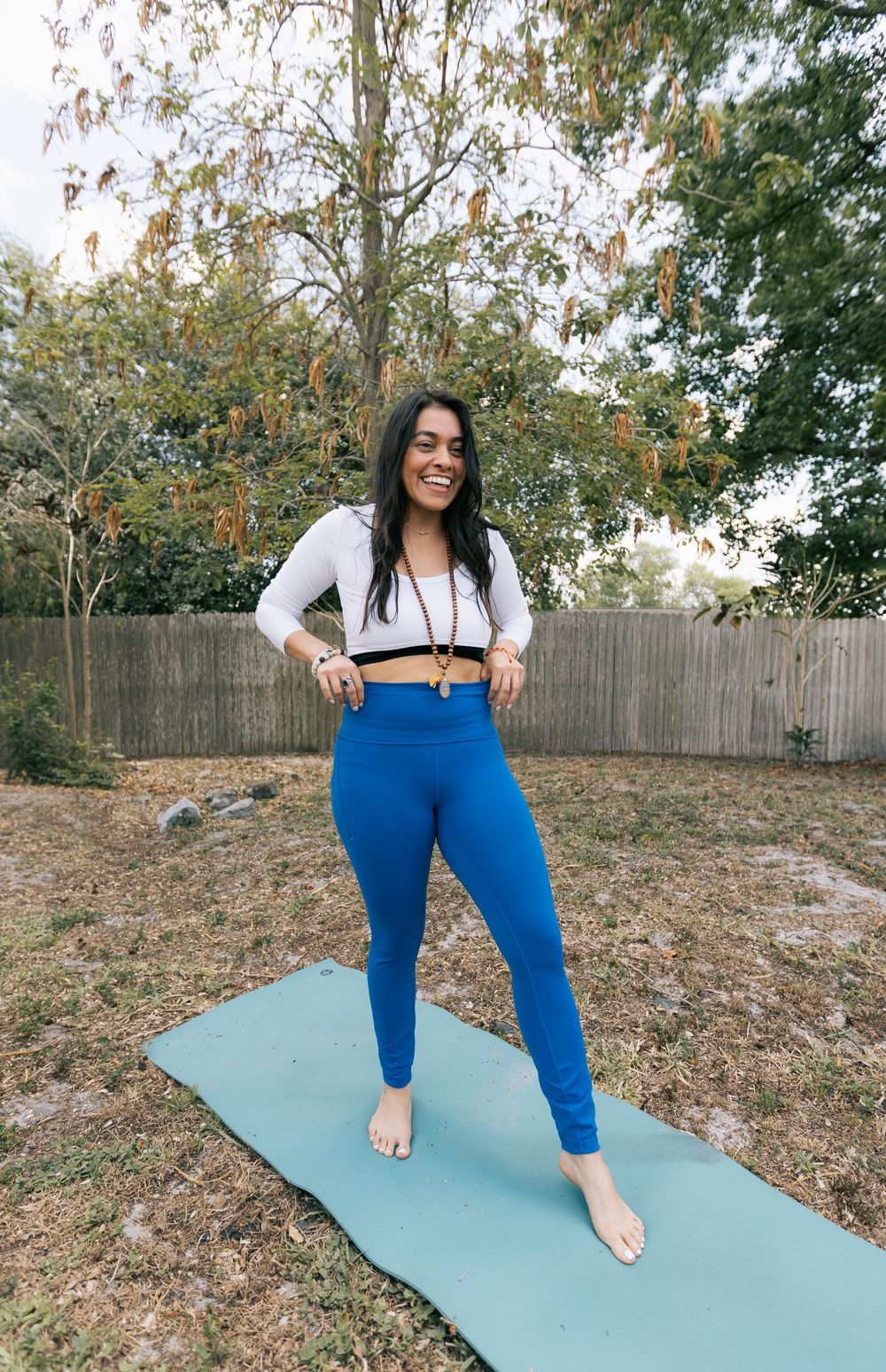 Smiling woman in blue leggings and white crop top standing barefoot on a teal yoga mat in a sunny backyard.