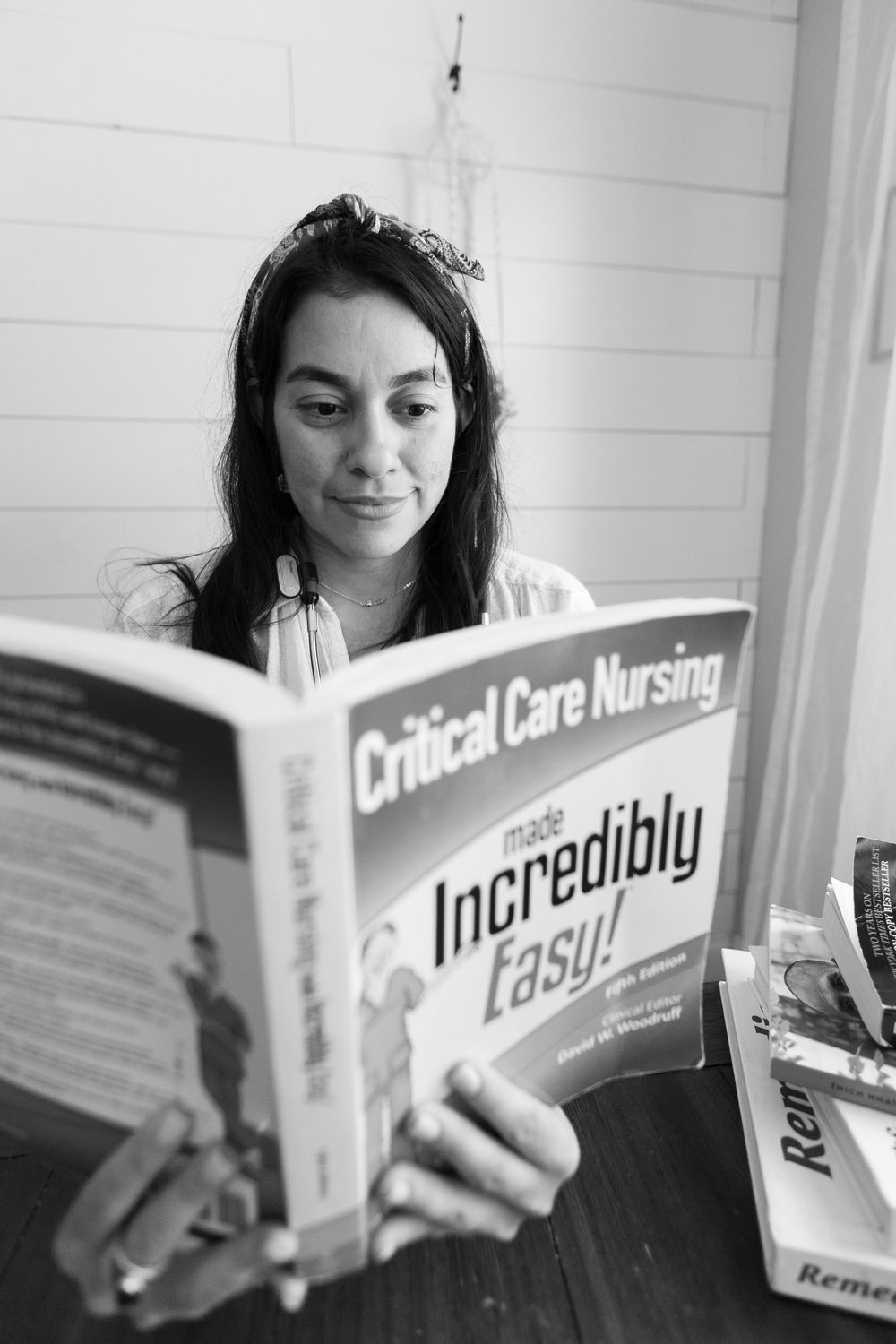 Black and white photo of a woman reading a Critical Care Nursing Made Incredibly Easy book at a table with stacked books.