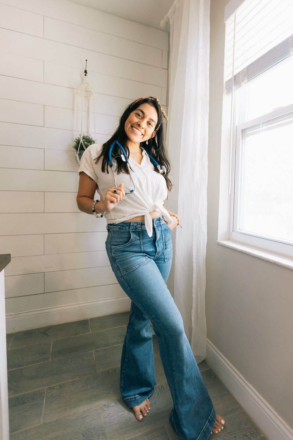 Smiling woman in flared jeans and a knotted white shirt with a blue bandana stands barefoot near a window.