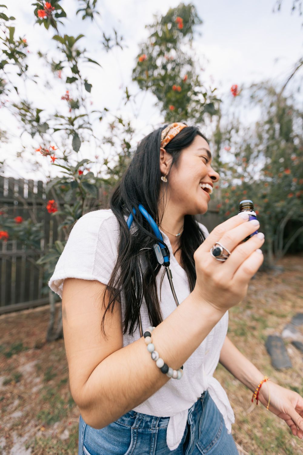 Laughing woman in a white tee and denim holds a small essential oil bottle outdoors near blooming red flowers.