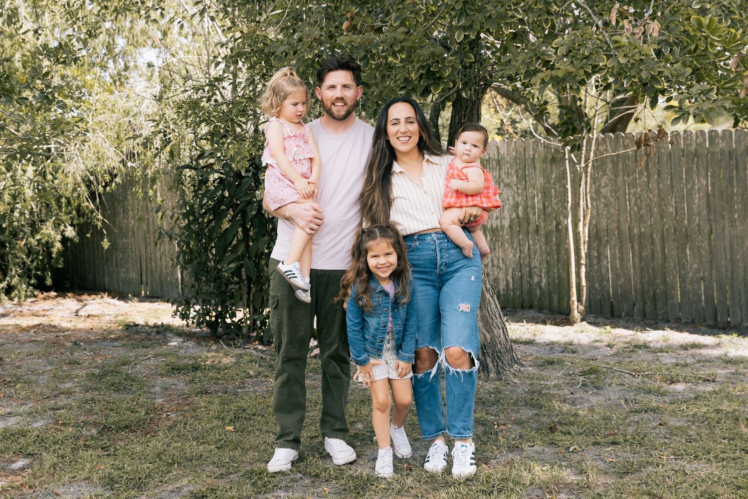 Smiling family of five, parents holding two toddlers with a young girl in front, poses in a sunny backyard by a wooden fence.