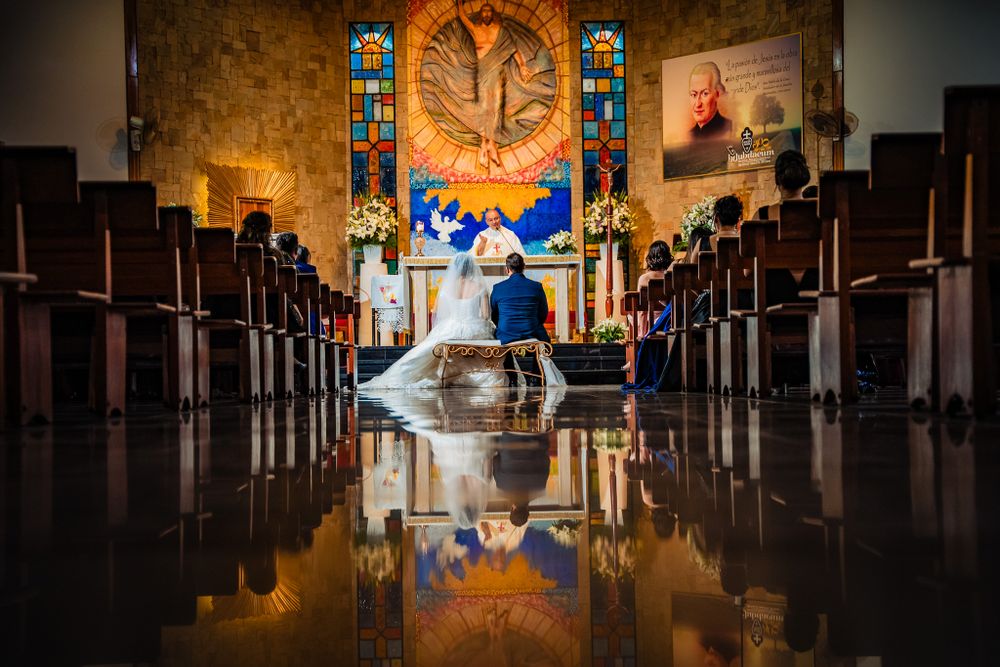 Bride and groom kneel at the altar in a beautifully decorated church, with a stunning reflection on the polished floor capturing the moment.