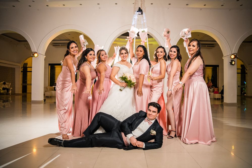 Bride and bridesmaids in elegant pink dresses pose playfully, as groom lounges confidently in front, creating fun and stylish wedding photo.