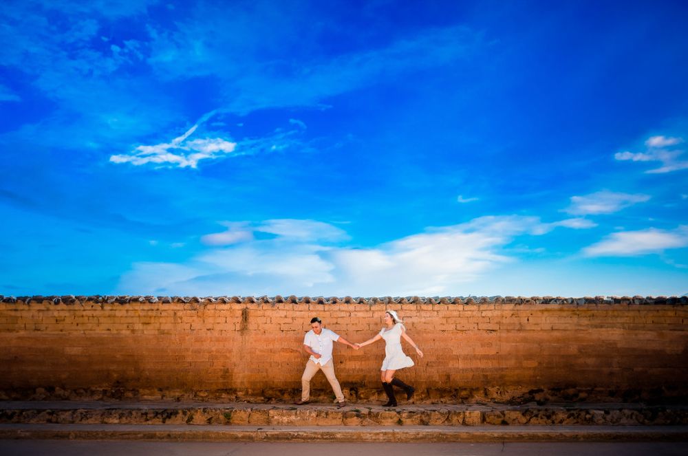 Joyful couple dances playfully in front of a rustic brick wall under a vibrant blue sky, creating a fun and romantic engagement moment.
