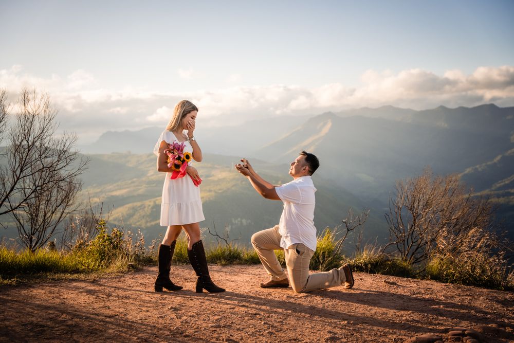 Romantic mountain-top proposal as a man kneels with a ring, surprising his partner holding a bouquet, with a breathtaking scenic backdrop.