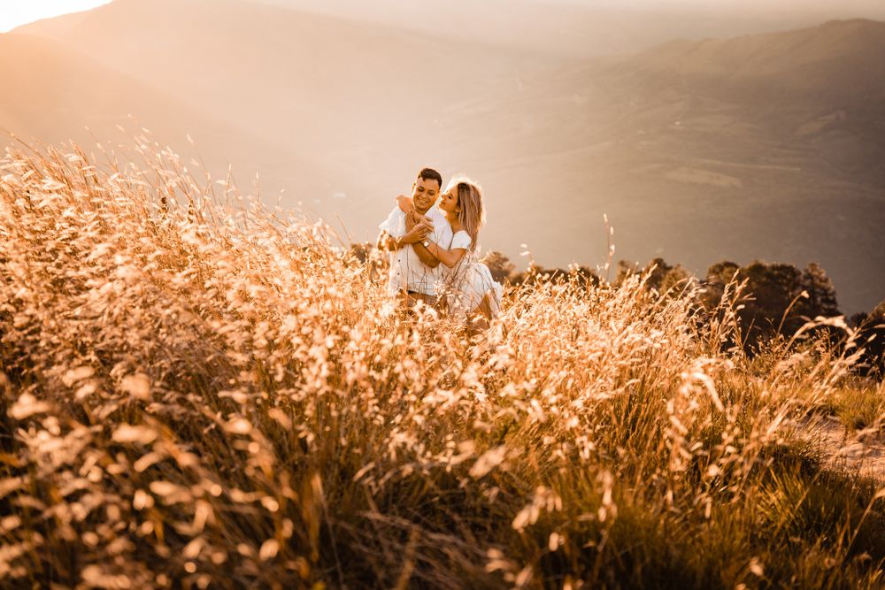 Romantic couple embraces in a golden field at sunset, surrounded by tall grass, with a breathtaking mountain landscape in the background.