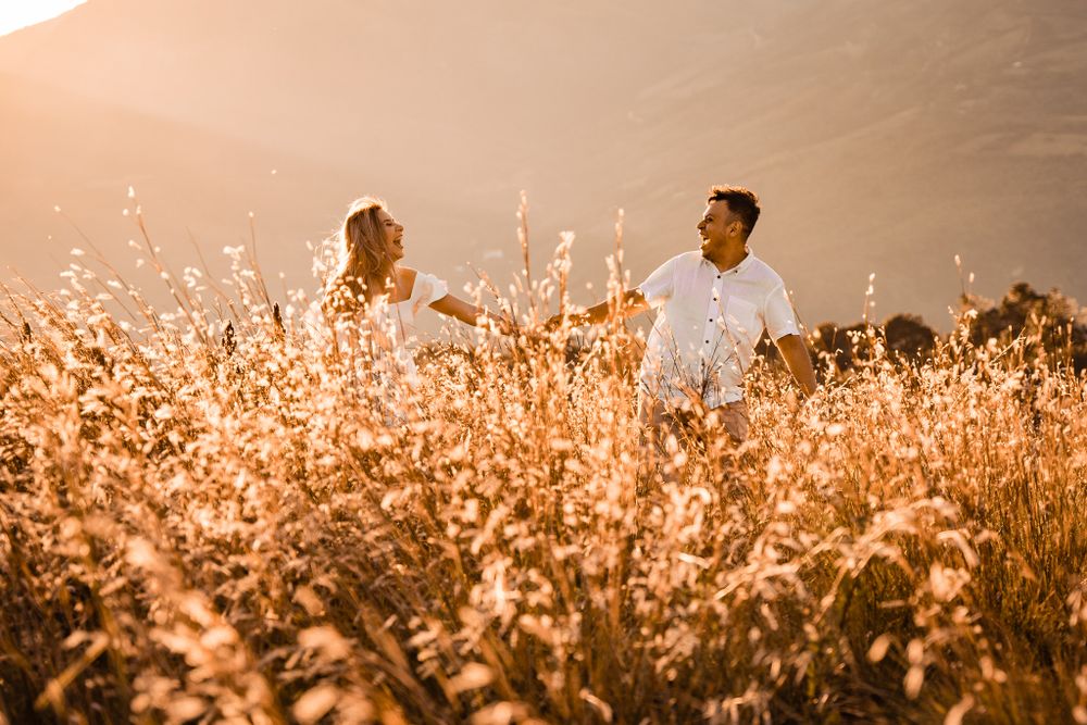 Romantic couple holding hands, walking through a golden field at sunset, surrounded by tall grass with mountain landscape in the background.