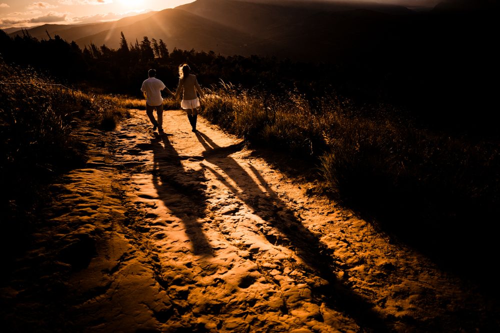 Romantic couple walks hand in hand along a sunlit trail at sunset, casting long dramatic shadows against a breathtaking mountain landscape.