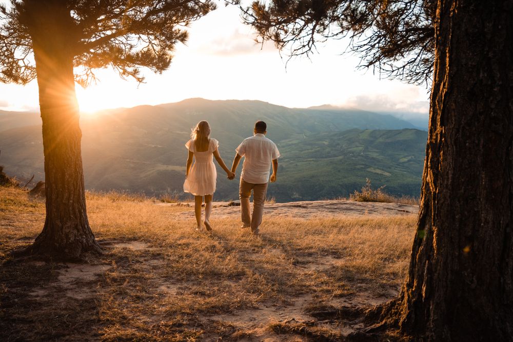 Romantic couple walks hand in hand through a golden landscape at sunset, framed by trees, overlooking a breathtaking mountain view.