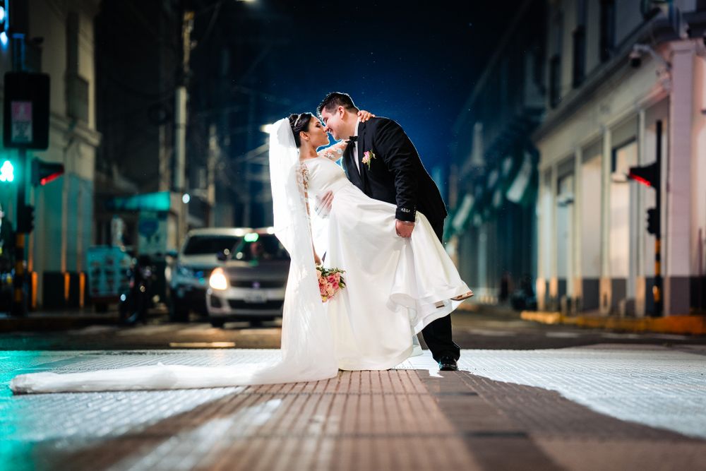 Newlywed couple shares a romantic dip kiss in the middle of a city street at night, illuminated by streetlights and passing traffic.