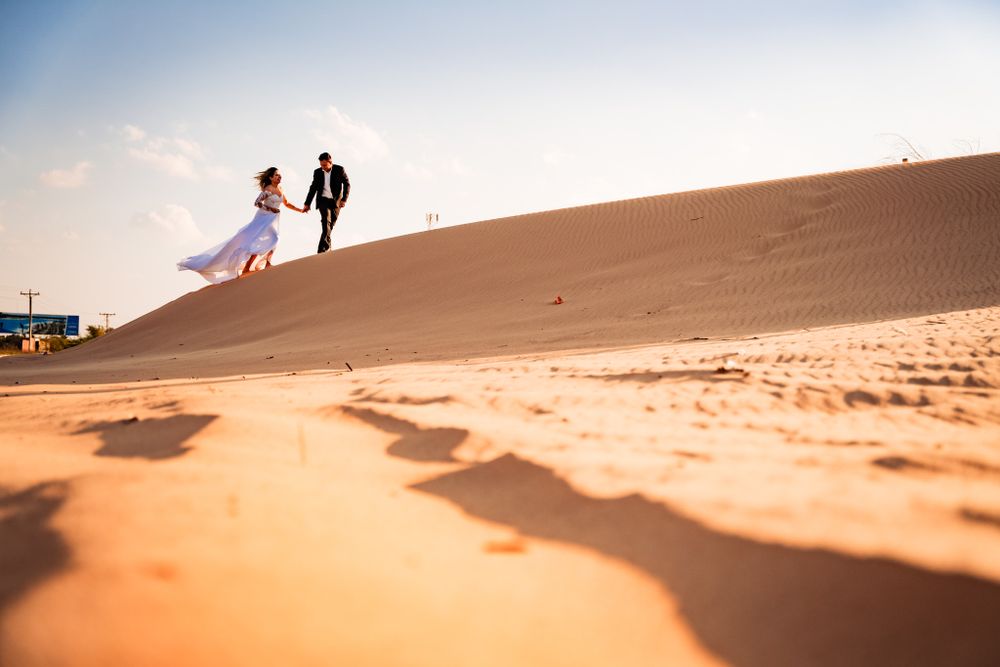 Newlywed couple joyfully runs hand in hand across sunlit sand dune, with bride’s dress flowing in the breeze, creating a dynamic moment.