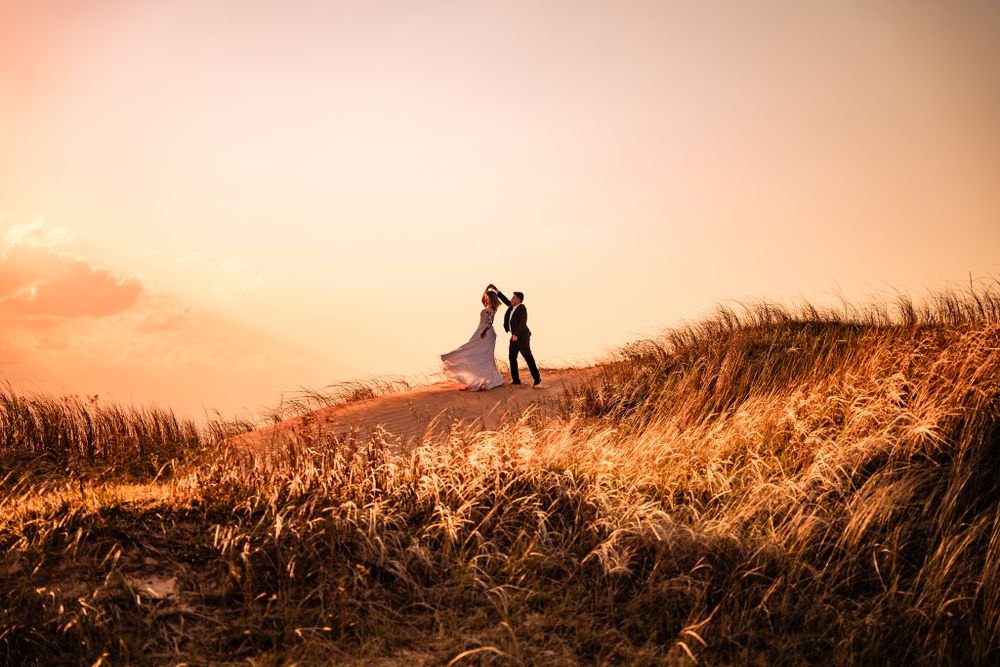 Newlywed couple shares romantic moment atop a golden sand dune at sunset, with bride’s dress flowing in the wind, creating a dreamy scene.