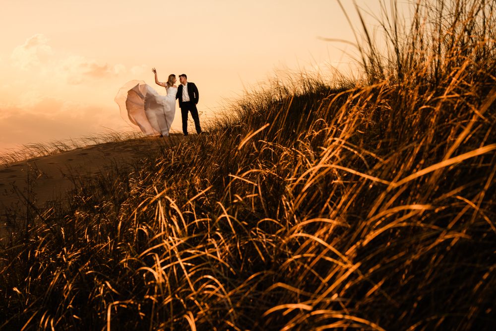 Newlywed couple stands atop a golden sand dune at sunset, with the bride’s veil flowing in the wind, creating a romantic and scenic moment.