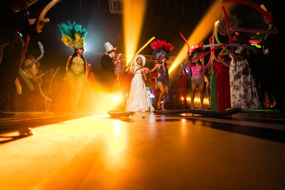 Bride and groom celebrate on vibrant dance floor, near performers in colorful costumes and dramatic lighting, creating a festive scene.