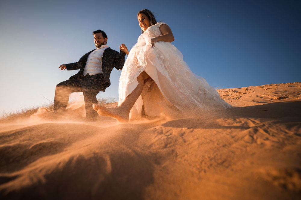 Newlywed couple joyfully descends a sunlit sand dune, kicking up golden sand, creating a dynamic and adventurous wedding moment.