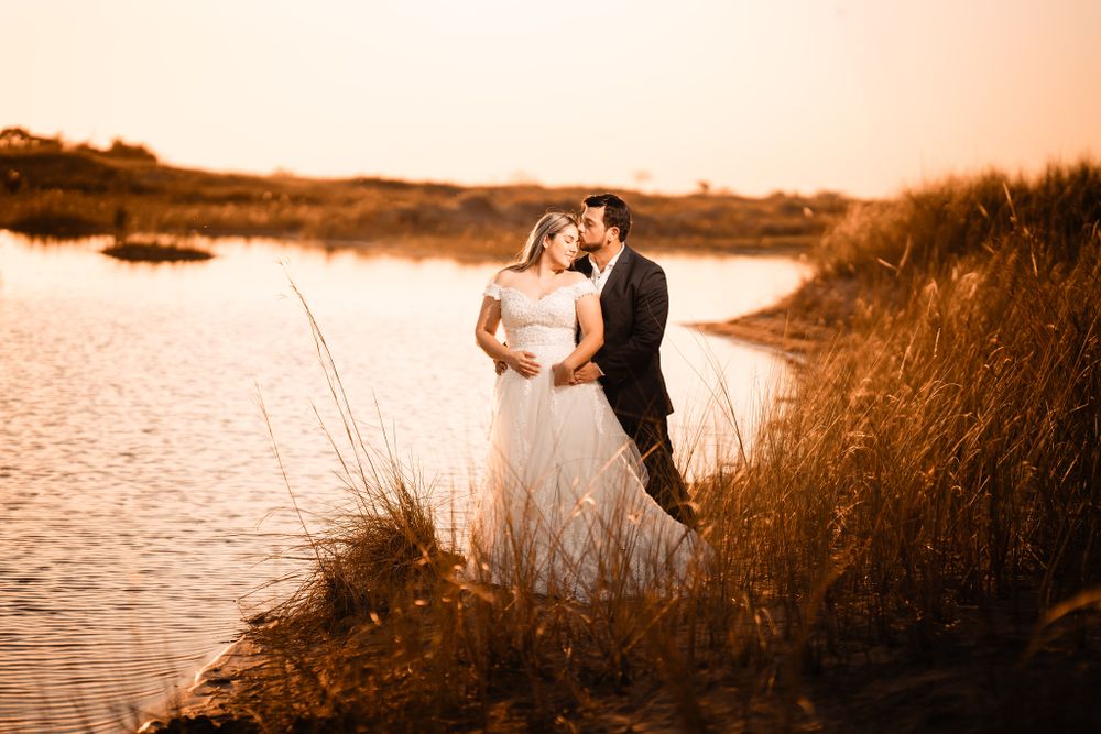 Bride and groom embrace by a peaceful lakeside at sunset, surrounded by golden grass, creating a romantic and serene wedding moment.