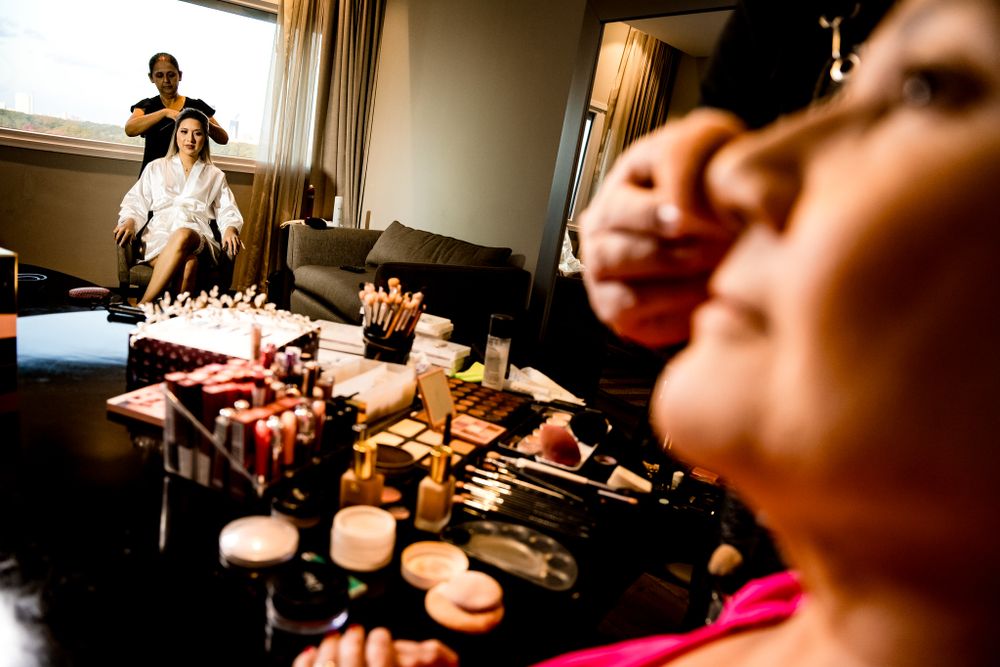 Bridal preparation with a makeup artist working in foreground, while bride gets her hair done in the background, capturing the excitement.