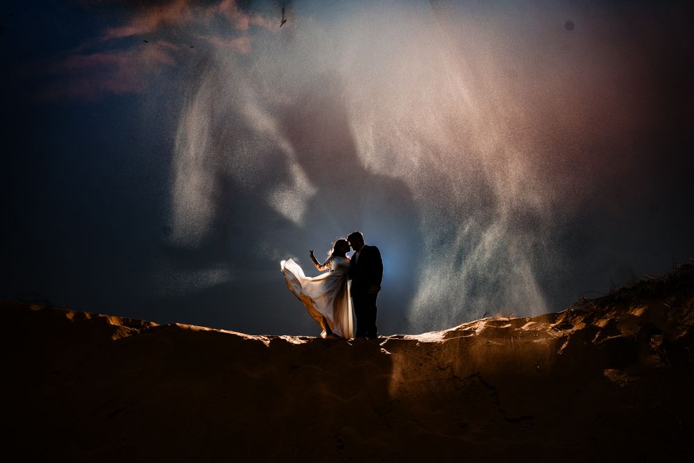Silhouetted newlywed couple embraces on a sand dune under a dramatic night sky, with flowing mist and light creating a magical atmosphere.