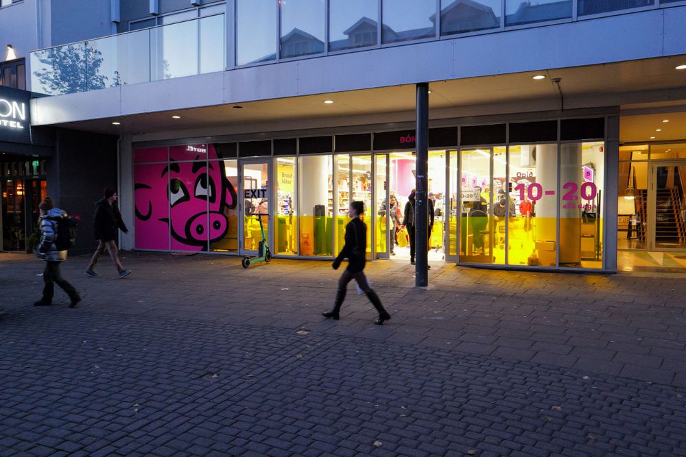 People walking past a brightly lit storefront with pink and yellow illuminated displays at dusk on a cobblestone street.