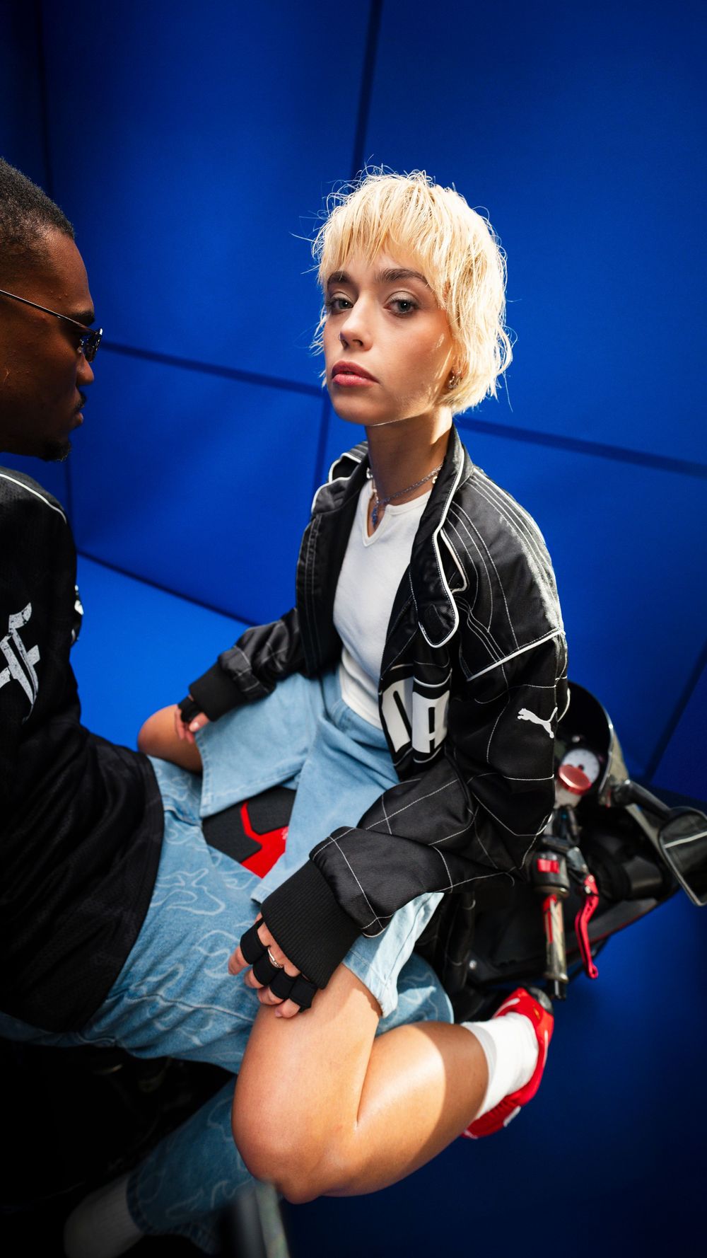 Fashion portrait of a person with short blonde hair wearing dark clothing in a bold blue studio environment