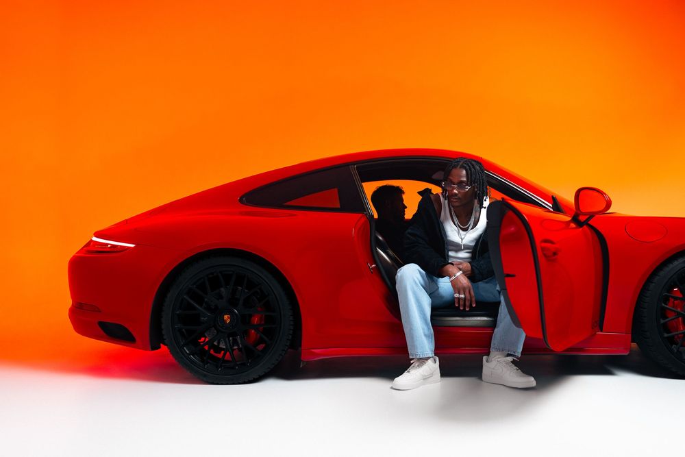 Fashion automotive photo of a man in casual sportswear sitting in front of a red Porsche against an orange background