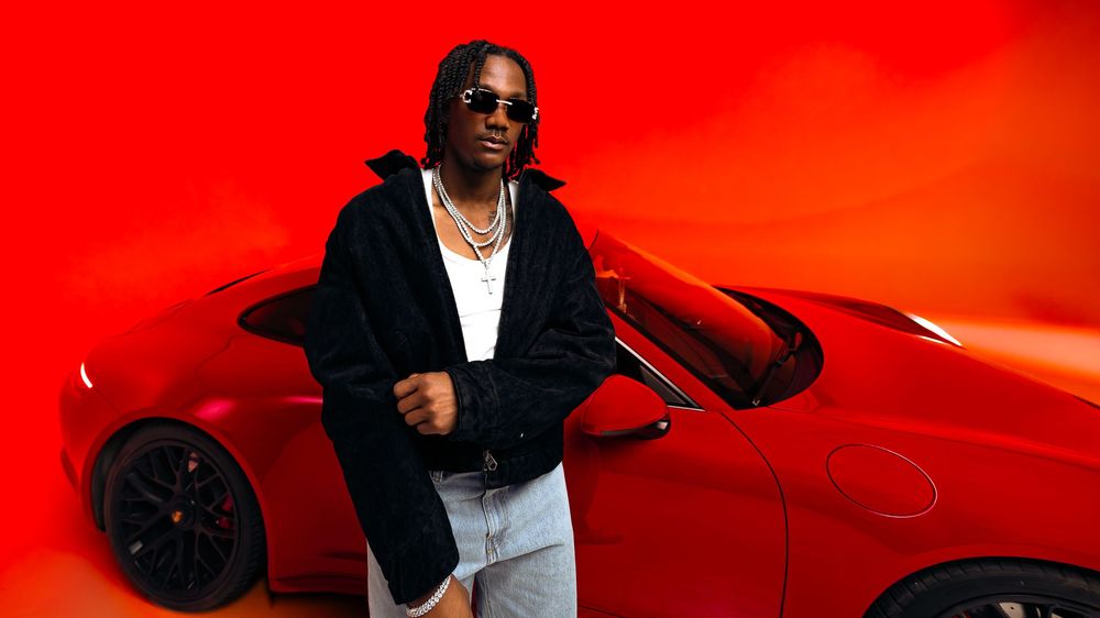 Fashion and automotive photo of a man in a black jacket leaning against a red Porsche on an intense red-orange background