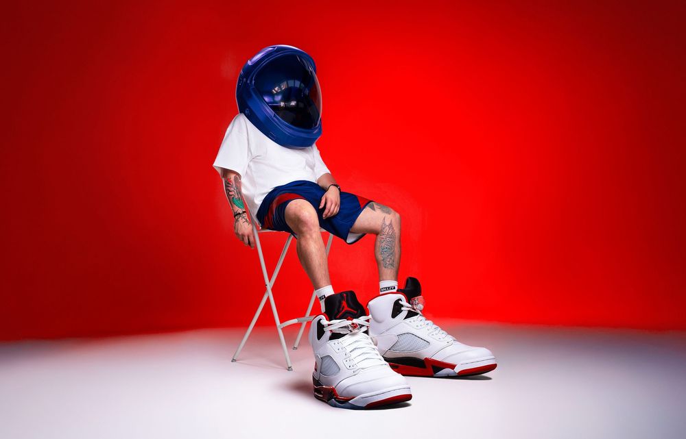 Sneaker campaign photo of a person in white sportswear and a blue bucket hat, seated on a folding chair against a vivid red background