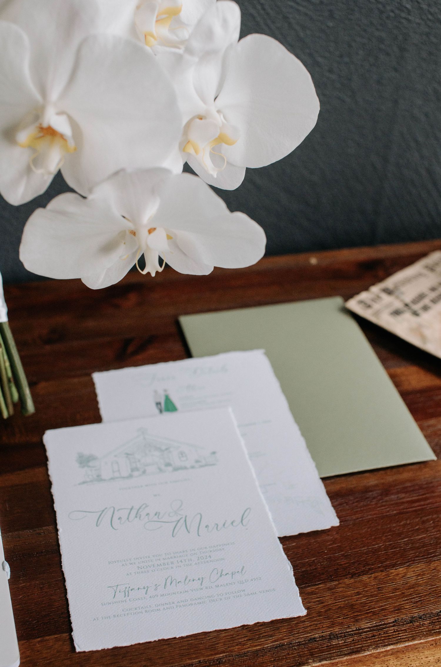 White orchid flowers and wedding invitation stationery displayed on a wooden table.
