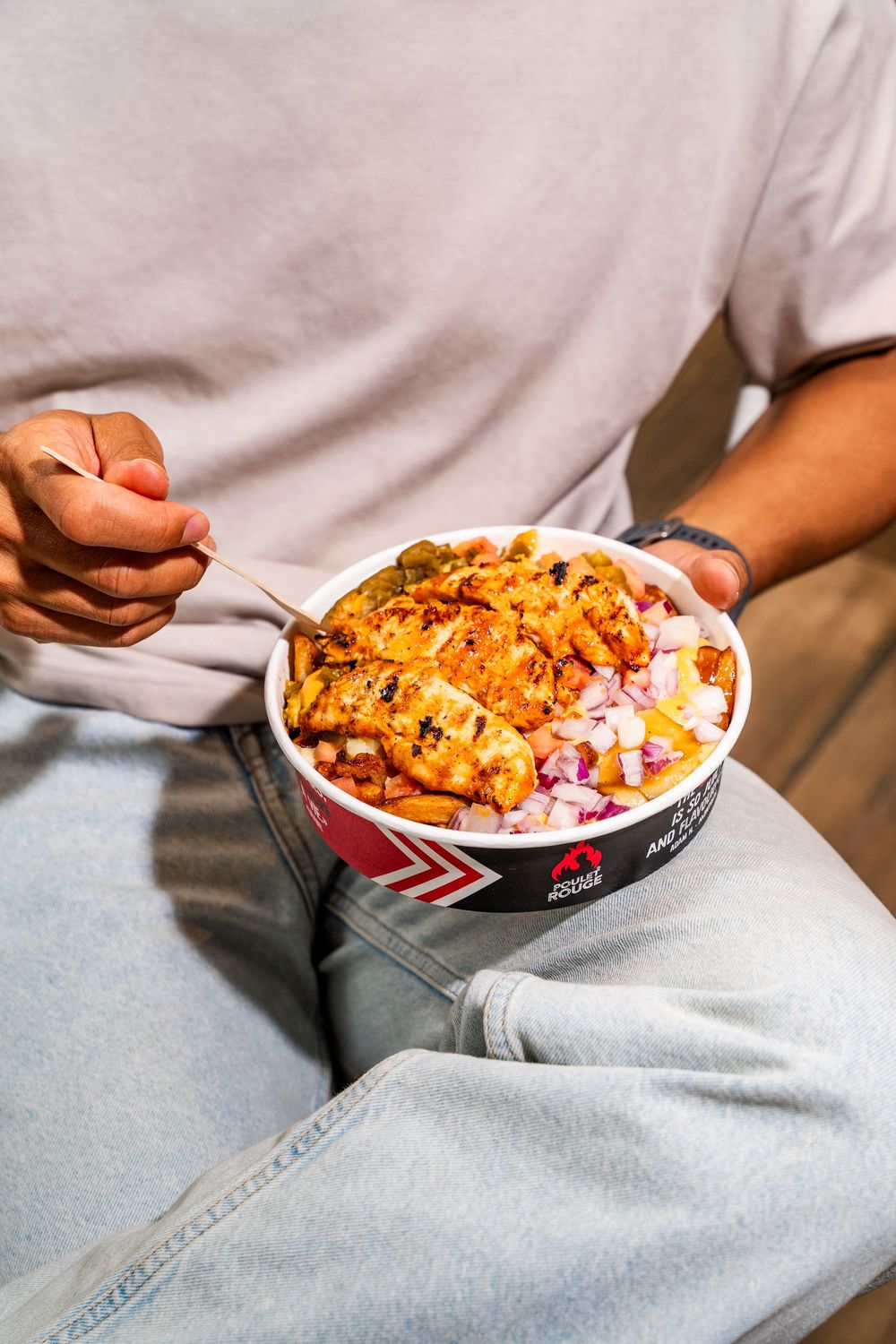 Person holding takeout container filled with spicy orange chicken and rice while sitting in casual denim attire.