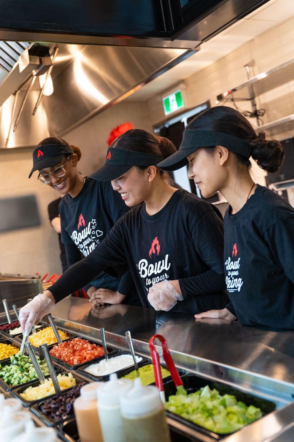 Restaurant workers in black uniforms prepare food at a salad bar station with fresh ingredients and toppings.