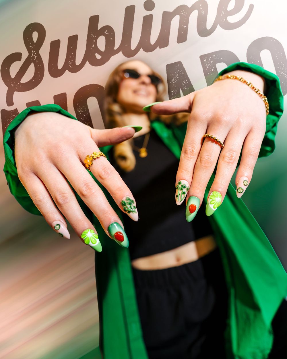 Close-up of hands with green and gold nail art designs wearing rings against a green fabric background.
