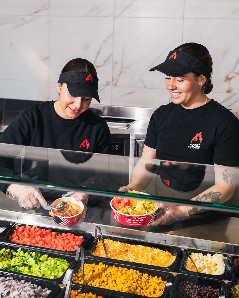 Restaurant workers in black uniforms and caps preparing fresh ingredients at a food service counter behind glass dividers.