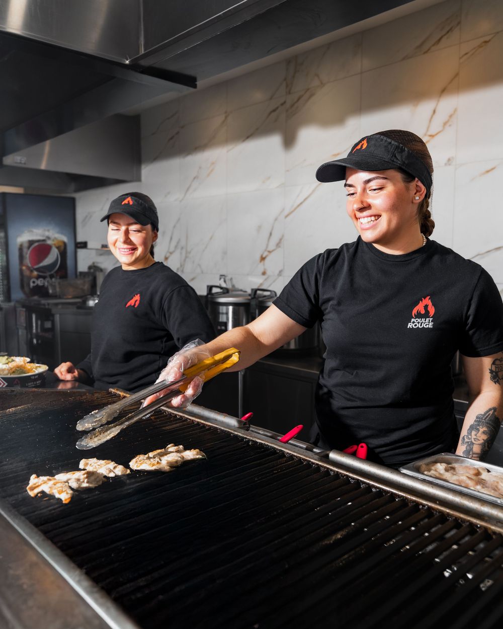 Restaurant workers in black uniforms and hats grilling food on a commercial flat top grill in a professional kitchen.