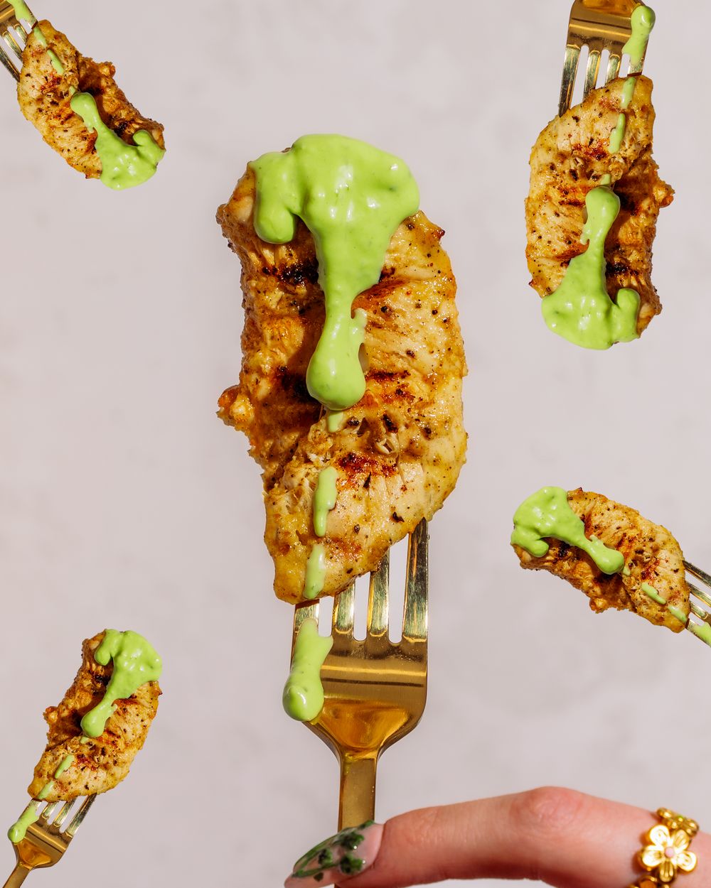 Grilled chicken skewers drizzled with bright green avocado sauce on silver forks against a white background.