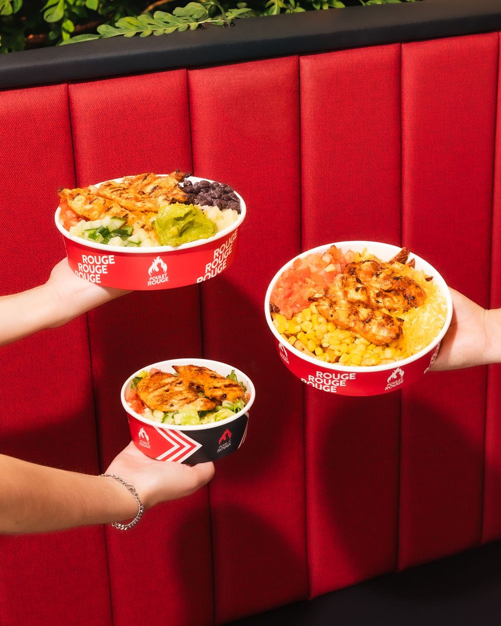 Three hands holding Rouge Rouge branded bowls filled with colorful rice dishes topped with grilled chicken and fresh ingredients.