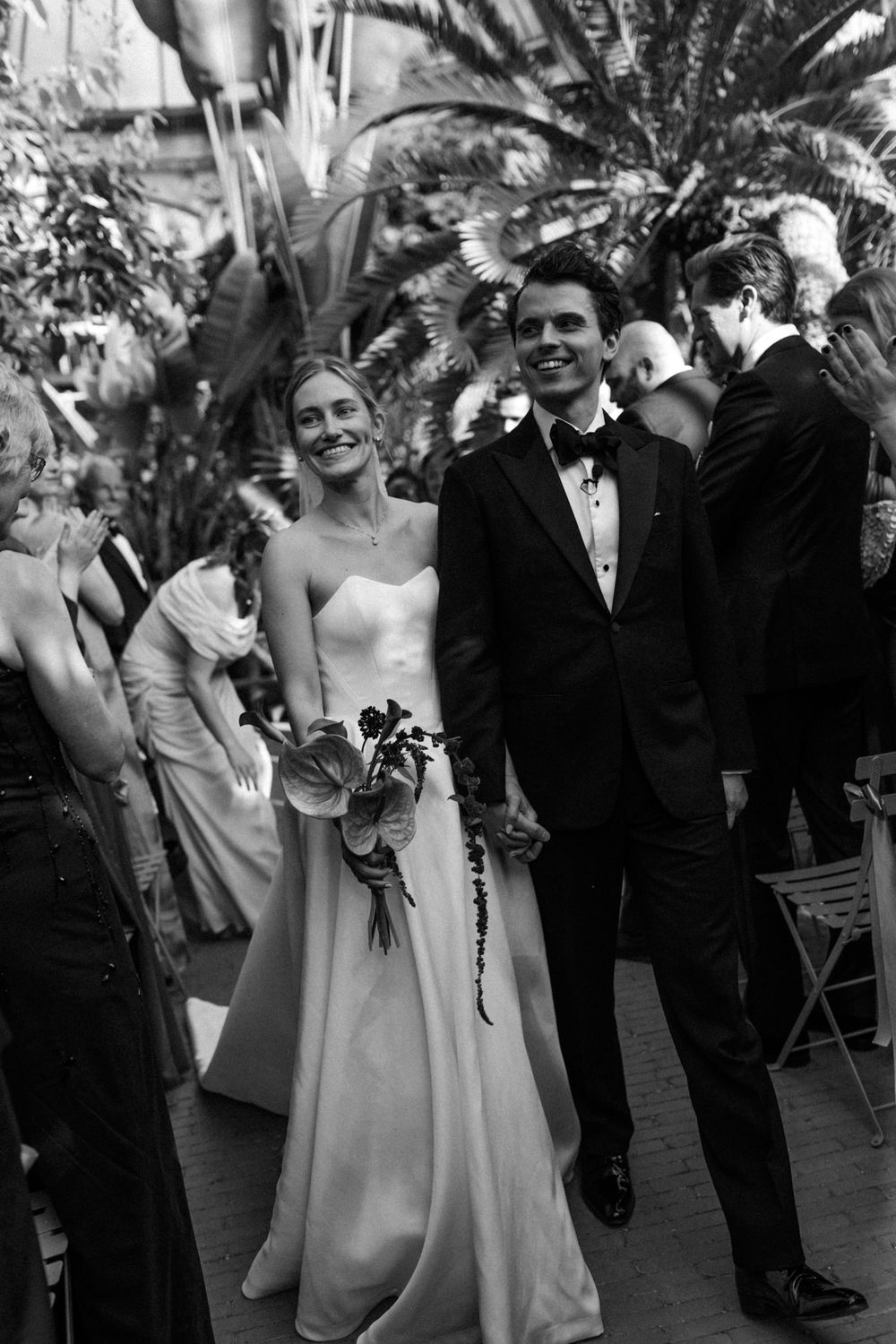 Black and white photo of a smiling bride in strapless gown holding bouquet walking with groom in tuxedo at wedding reception.