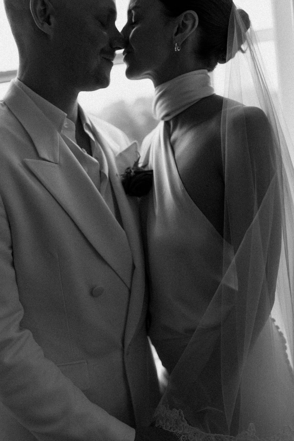 Black and white photo of a bride and groom sharing a romantic kiss, bride wearing backless gown with veil.