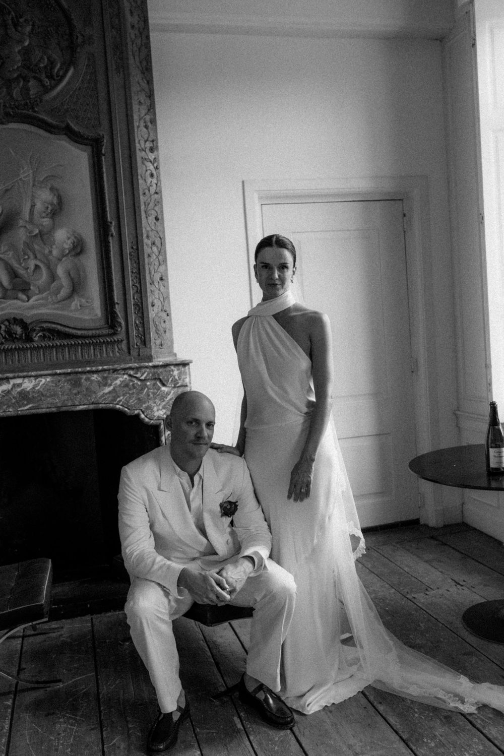 Black and white photo of a bride in elegant halter gown and groom in white suit posing in a chic vintage room.