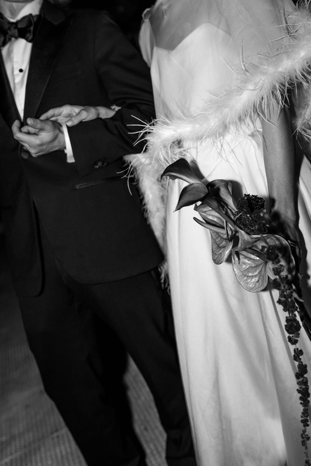 Black and white photo of bride in white gown with feather wrap holding bouquet beside groom in tuxedo.
