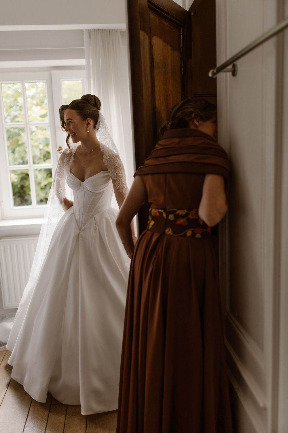 Bride in white ballgown wedding dress smiles near window while bridesmaid in brown satin gown stands nearby.